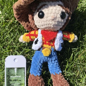 Puede incluir: Mu&ntilde;eco de ganchillo hecho a mano de un personaje de vaquero, con sombrero marr&oacute;n, chaleco blanco y pantalones azules. El mu&ntilde;eco est&aacute; colocado sobre hierba verde junto a un frasco de desinfectante de manos. El dise&ntilde;o del mu&ntilde;eco est&aacute; inspirado en una pel&iacute;cula de animaci&oacute;n popular.