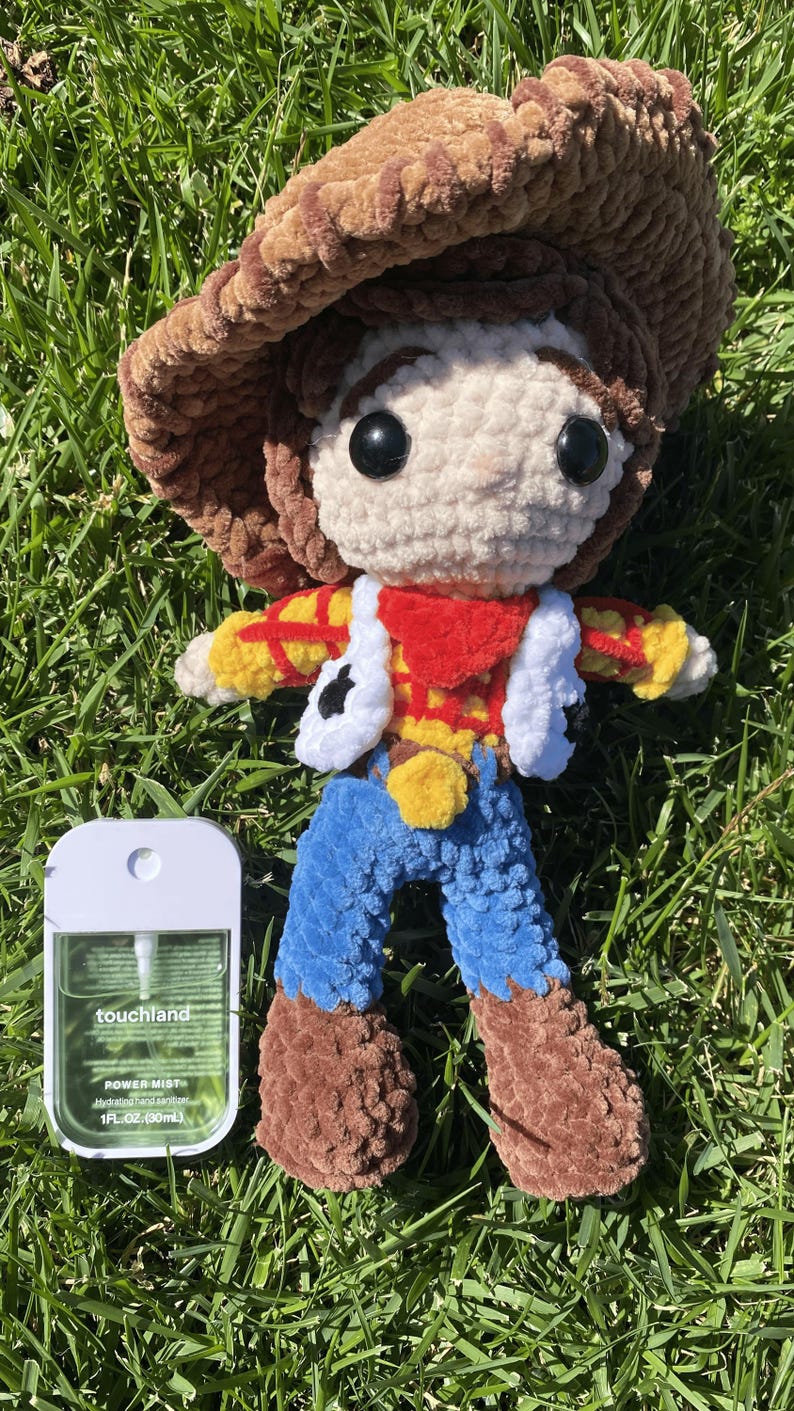 Puede incluir: Mu&ntilde;eco de ganchillo hecho a mano de un personaje de vaquero, con sombrero marr&oacute;n, chaleco blanco y pantalones azules. El mu&ntilde;eco est&aacute; colocado sobre hierba verde junto a un frasco de desinfectante de manos. El dise&ntilde;o del mu&ntilde;eco est&aacute; inspirado en una pel&iacute;cula de animaci&oacute;n popular.
