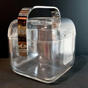 May include: A clear, square ice bucket with a chrome handle. The bucket has a rounded top and a textured panel on one side. The handle arches over the top. The ice bucket is designed for serving beverages, ideal for parties or gatherings.