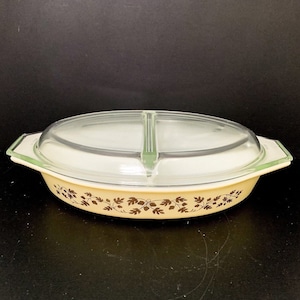 May include: Cream-coloured, oval divided casserole dish with a clear glass lid. The dish features a brown leaf pattern. The lid has a green handle and trim. A vintage kitchenware item for serving food.