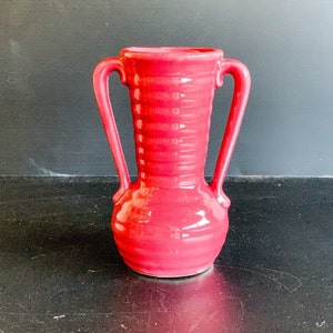 Puede incluir: Un jarrón de cerámica rojo con dos asas. El jarrón tiene un diseño acanalado y un acabado brillante.