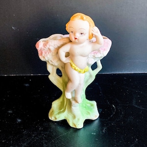 Puede incluir: Figura de porcelana vintage de una figura querubínica posando junto a un jarrón con forma de árbol. La figura tiene cabello rubio, una banda amarilla en la cintura y se encuentra sobre un fondo de tonos rosas y verdes. El jarrón mide unos 13 cm de alto.