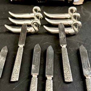 May include: A collection of six silver butter knives with decorative handles and swan-shaped rests. The knives feature a floral design on the blades and handles. The packaging reads "Set of 6 pieces" and "Ensemble de 6 morceaux."