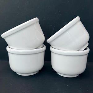 Op de afbeelding: Vier witte keramische ramekins, in paren gestapeld. Elke ramekin heeft een afgeronde basis en een licht uitlopende rand. De ramekins zijn geplaatst tegen een donkere achtergrond, wat hun strakke, eenvoudige ontwerp benadrukt. Ideaal voor bakken en serveren.