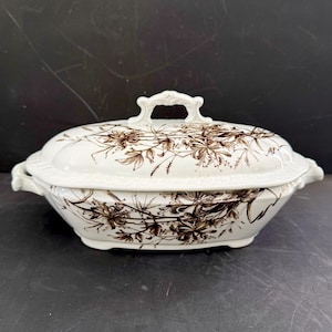 May include: A white ceramic serving dish with a lid, featuring a brown floral pattern. The dish has a handle on the lid and two side handles. The design is a classic, vintage style, suitable for serving food.