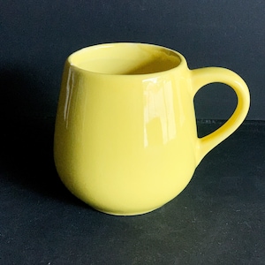 May include: A glossy, yellow ceramic mug with a rounded body and a curved handle. The mug has a smooth, reflective surface and is designed for holding hot or cold beverages. The mug is a solid, bright yellow color.