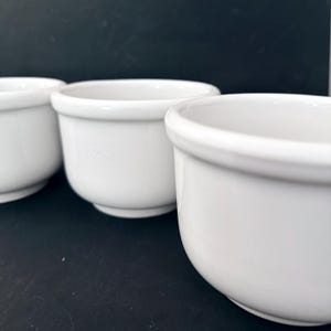Puede incluir: Tres cuencos de cer&aacute;mica blanca con bordes redondeados. Los cuencos est&aacute;n dispuestos en fila, con el cuenco m&aacute;s cercano enfocado. Los cuencos tienen un dise&ntilde;o simple y cl&aacute;sico, adecuado para servir sopas, postres u otros platos.