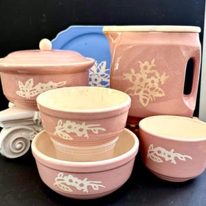 Pode incluir: Uma coleção de utensílios de cozinha de cerâmica rosa vintage. Inclui uma assadeira com tampa, tigelas, um jarro e um prato azul. Cada peça apresenta um design floral branco. As tigelas estão empilhadas.
