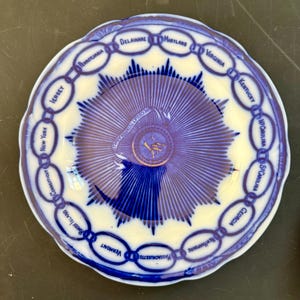 May include: A vintage blue and white decorative plate featuring a central design with radiating lines and the text "Deus Et Humanum Agit". The plate's rim is adorned with the names of U.S. states in a chain-like pattern.