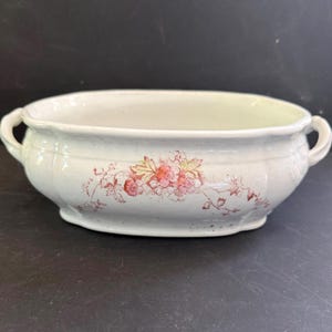 May include: An oval-shaped white ceramic serving bowl featuring a delicate pink and orange floral transfer pattern on the front. The bowl has a subtle crackle glaze finish, a scalloped rim, and two small curved handles on opposite sides.
