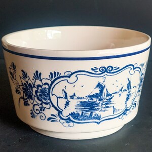 Puede incluir: Un cuenco de cer&aacute;mica blanco con un borde azul y un dise&ntilde;o decorativo azul. El dise&ntilde;o incluye motivos florales y una escena de paisaje enmarcada con barcos y edificios. El cuenco tiene una forma ligeramente acampanada.