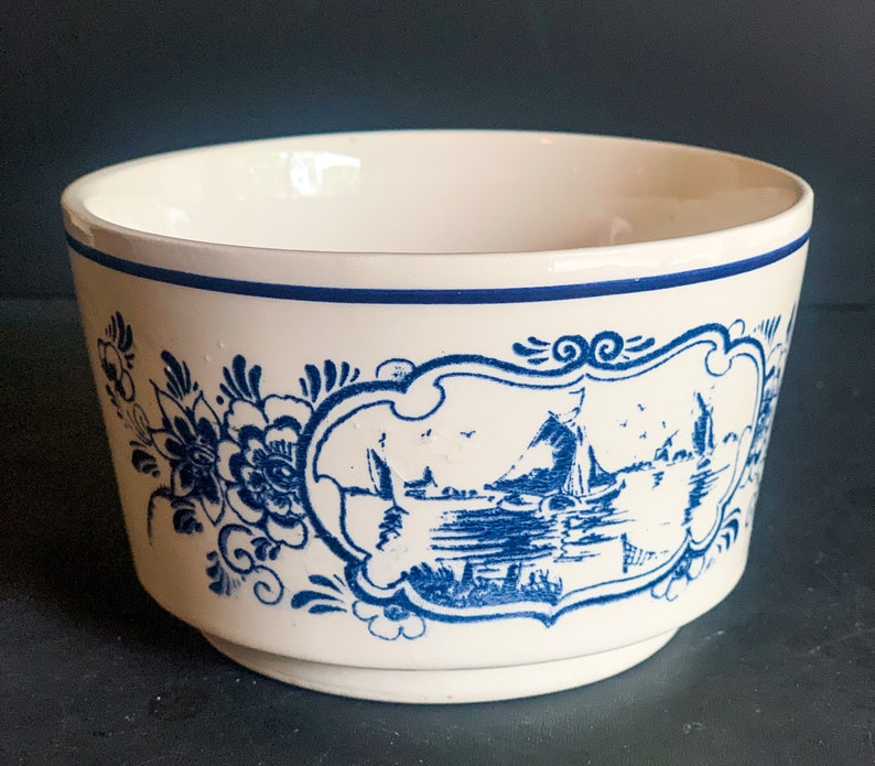 Puede incluir: Un cuenco de cer&aacute;mica blanco con un borde azul y un dise&ntilde;o decorativo azul. El dise&ntilde;o incluye motivos florales y una escena de paisaje enmarcada con barcos y edificios. El cuenco tiene una forma ligeramente acampanada.