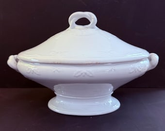 May include: A white ceramic tureen with a matching lid, featuring a glossy finish and embossed leaf patterns along the rim and base. The vessel includes two side handles and a central loop handle on the lid, resting on a pedestal base.