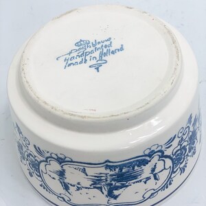 Puede incluir: Un cuenco de cer&aacute;mica blanco con dise&ntilde;os florales y paisaj&iacute;sticos azules. El cuenco tiene forma redondeada y un borde ancho. La base del cuenco tiene el texto "Deft Blauw Handpainted made in Holland".