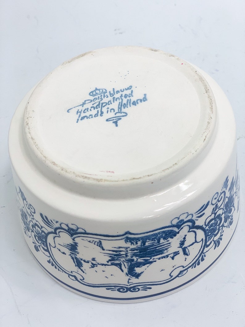 Puede incluir: Un cuenco de cer&aacute;mica blanco con dise&ntilde;os florales y paisaj&iacute;sticos azules. El cuenco tiene forma redondeada y un borde ancho. La base del cuenco tiene el texto "Deft Blauw Handpainted made in Holland".