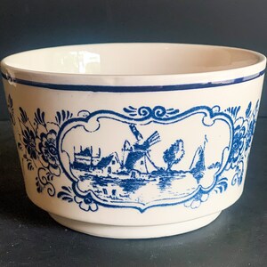 Puede incluir: Cuenco de cer&aacute;mica color crema con una escena azul de molino de viento holand&eacute;s. El cuenco presenta un borde azul y un dise&ntilde;o decorativo floral y de volutas azules. La imagen central representa un molino de viento, casas y un barco.