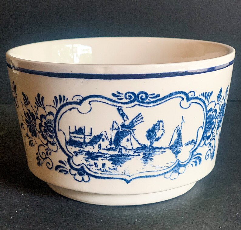 Puede incluir: Cuenco de cer&aacute;mica color crema con una escena azul de molino de viento holand&eacute;s. El cuenco presenta un borde azul y un dise&ntilde;o decorativo floral y de volutas azules. La imagen central representa un molino de viento, casas y un barco.
