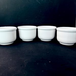 Puede incluir: Cuatro cuencos peque&ntilde;os de cer&aacute;mica blanca con forma redondeada. Los cuencos tienen un dise&ntilde;o sencillo y cl&aacute;sico y est&aacute;n dispuestos en fila sobre un fondo oscuro. Probablemente sean para servir salsas o porciones peque&ntilde;as. Cada cuenco mide unos 10 cm de di&aacute;metro.