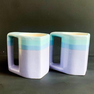 Peut inclure: Deux tasses en céramique avec un glaçage bleu et violet. Les tasses ont une forme carrée et une poignée sur le côté.