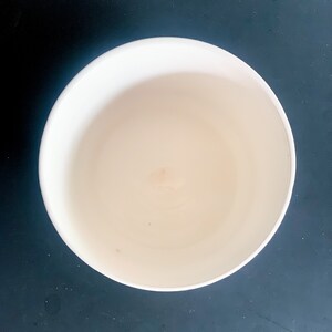 Puede incluir: Vista superior de un cuenco de cer&aacute;mica blanco roto vac&iacute;o. El cuenco tiene una forma redondeada con un interior liso y un exterior ligeramente texturizado. El cuenco est&aacute; sobre un fondo oscuro.