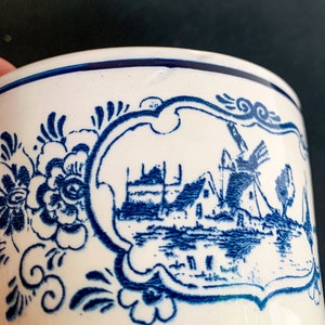 Puede incluir: Primer plano de una taza de cer&aacute;mica blanca con un dise&ntilde;o de molino de viento holand&eacute;s azul. La taza presenta un patr&oacute;n floral azul y una escena de paisaje enmarcada. El borde est&aacute; acentuado con una franja azul.