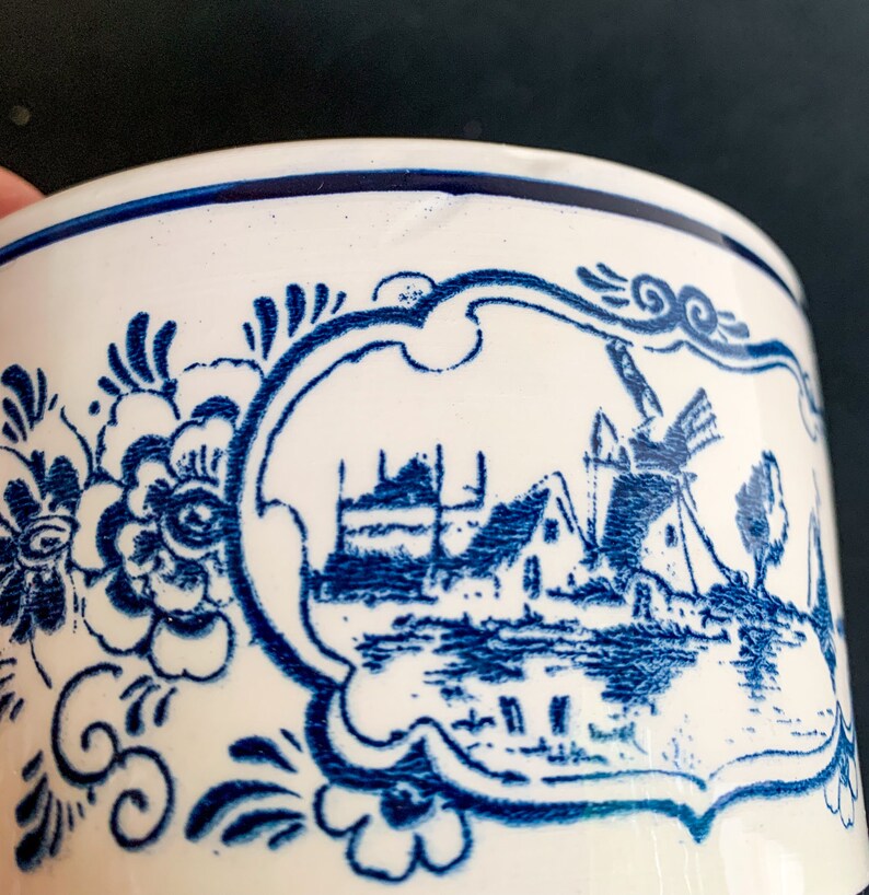 Puede incluir: Primer plano de una taza de cer&aacute;mica blanca con un dise&ntilde;o de molino de viento holand&eacute;s azul. La taza presenta un patr&oacute;n floral azul y una escena de paisaje enmarcada. El borde est&aacute; acentuado con una franja azul.