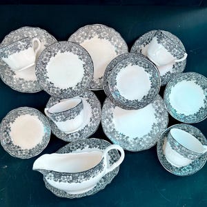 May include: A vintage china set featuring a white and grey floral design. The collection includes teacups, saucers, plates, and a gravy boat. The floral pattern decorates the rims of the pieces. The china is displayed on a dark surface.