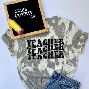 May include: A gray and white tie-dye t-shirt with the word "TEACHER" repeated three times in black, retro-style lettering. The shirt is laying on a white surface with a pair of blue jeans and a small black letter board with the words "GOLDEN GRATITUDE CO."