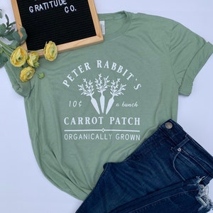 Puede incluir: Una camiseta verde salvia con un gráfico blanco de Peter Rabbit's Carrot Patch. El gráfico incluye un manojo de zanahorias con hojas y el texto "10¢ un manojo, Carrot Patch, Cultivado orgánicamente".