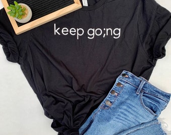 May include: Black t-shirt with the text "keep going" in white lettering. The shirt is laid flat on a white surface with a pair of blue jeans and a letter board that says "Golden Gratitude Co."