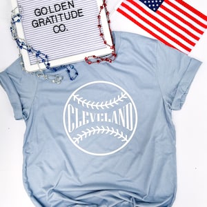 May include: A light blue t-shirt with a white baseball graphic that says "CLEVELAND". The shirt is on a white surface with a letter board that says "GOLDEN GRATITUDE CO.", a small American flag, and red and blue beaded necklaces.