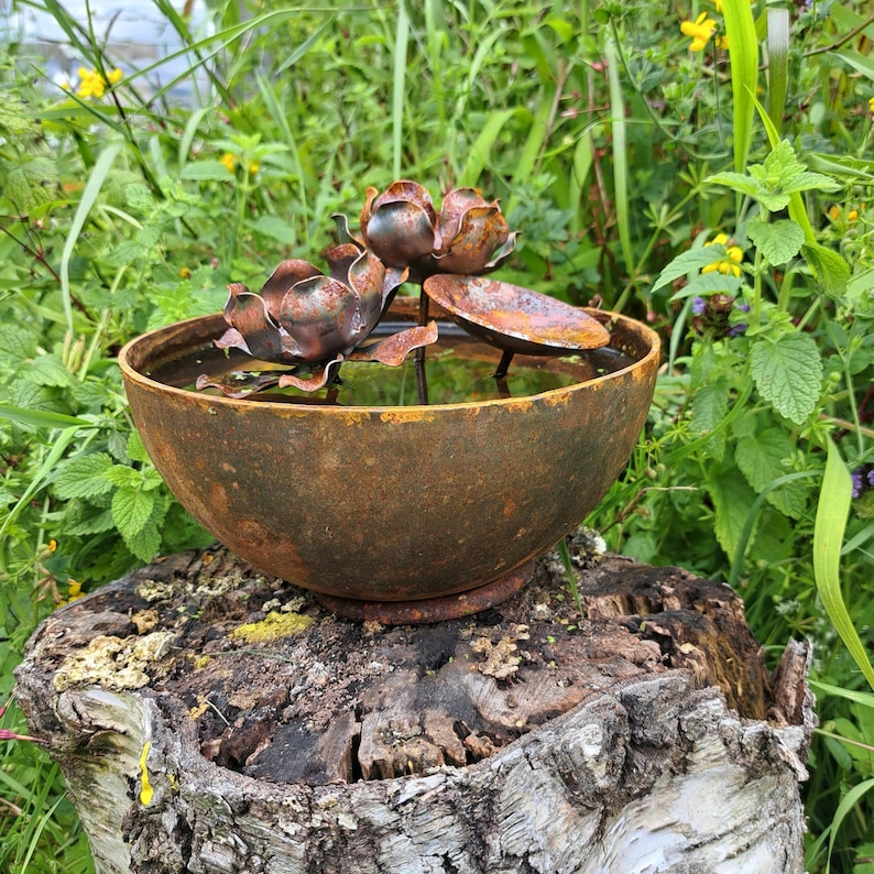 May include: A rustic, metal water fountain with a bowl-shaped basin and decorative lotus flowers. The fountain has a weathered, brown finish and sits atop a tree stump, surrounded by green foliage. The water feature adds a natural element to any garden.