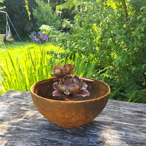 May include: A weathered, rusty metal bowl with a decorative floral sculpture inside. The sculpture features multiple bronze-colored flower heads. The bowl sits on a wooden surface, with a lush green garden in the background.