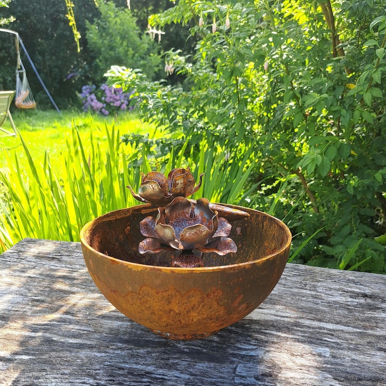 May include: A weathered, rusty metal bowl with a decorative floral sculpture inside. The sculpture features multiple bronze-colored flower heads. The bowl sits on a wooden surface, with a lush green garden in the background.