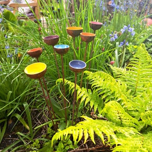 May include: A collection of colorful, handcrafted garden art pieces. Each piece features a small, bowl-shaped top in shades of yellow, orange, blue, and red, mounted on a slender, brown metal stem. The art is set amidst lush green foliage.