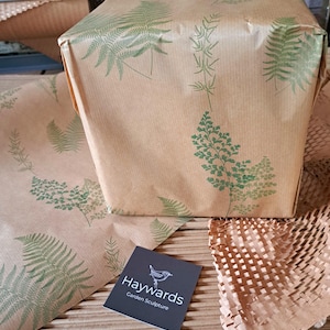 May include: A wrapped package and wrapping paper with a fern leaf pattern in green on brown paper. A business card with the text "Haywards Garden Sculpture" is in the foreground. The package is wrapped in brown paper with green fern leaf designs.