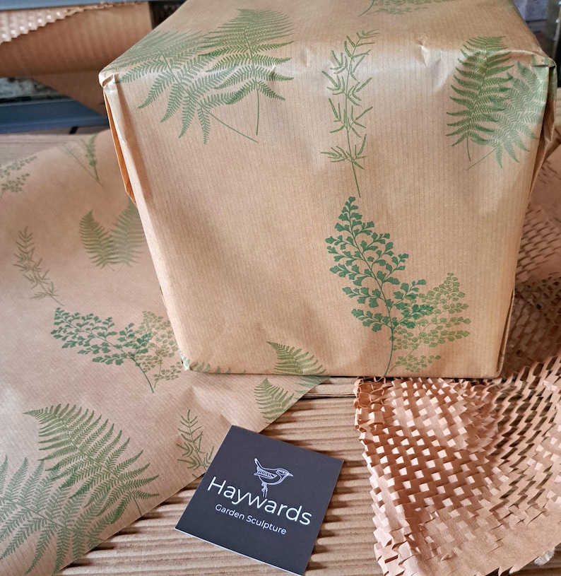 May include: A wrapped package and wrapping paper with a fern leaf pattern in green on brown paper. A business card with the text "Haywards Garden Sculpture" is in the foreground. The package is wrapped in brown paper with green fern leaf designs.