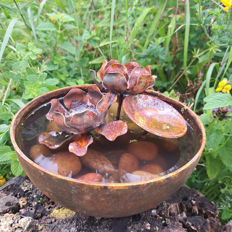 May include: A rustic, metal water feature with a lotus flower design. The bowl is filled with water and small stones. The lotus flowers are a rusty brown color, and the bowl has a weathered appearance. The water feature is set outdoors among green foliage.