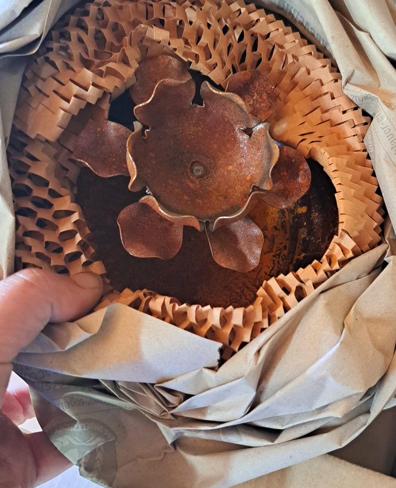 May include: A rusty brown metal flower sculpture, partially visible, nestled within protective brown paper packaging. The sculpture has multiple petals and a central circular element. The packaging includes honeycomb-style paper and folded brown paper.