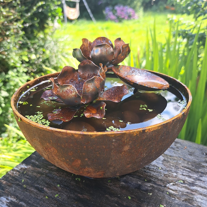 May include: A rustic, metal water bowl with a lotus flower sculpture. The bowl is filled with water and small green plants. The lotus flower is made of weathered metal, with brown and copper tones. The bowl sits on a wooden surface.