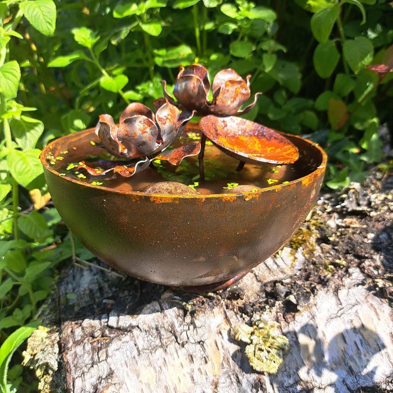 May include: A rustic, metal water fountain with a lotus flower design. The bowl-shaped fountain is filled with water and small green plants. The lotus flowers are made of metal and have a weathered, brown finish. The fountain sits on a tree stump.
