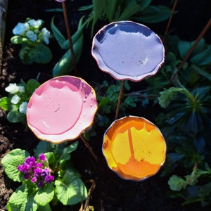 May include: Three colorful metal flower garden stakes. The flowers are in shades of pink, purple, and yellow, with a wavy edge. The stakes are in a garden bed with green foliage and other flowers.