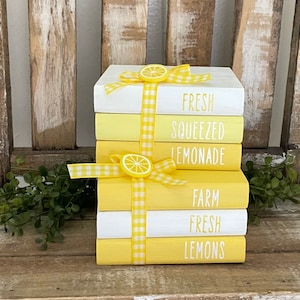 Puede incluir: Una pila de cinco libros pintados de amarillo y blanco con una cinta de cuadros amarillos y blancos atada alrededor. Los libros están decorados con una rodaja de limón y el texto "Fresh Squeezed Lemonade", "Farm Fresh Lemons".
