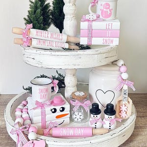 Può includere: Un vassoio a due livelli bianco decorato con un tema invernale. Il vassoio presenta figurine di pupazzo di neve rosa e bianco, una tazza rosa e bianca che dice "Snow Day" e un mattarello rosa e bianco che dice "Snow Day". Il vassoio ha anche un barattolo di ceramica bianco con un cuore nero e una tazza di ceramica bianca con un design di pupazzo di neve rosa. Il vassoio è decorato con un nastro a quadri rosa e bianco e perline bianche.