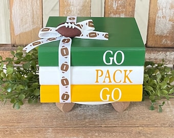 May include: A set of three stacked wooden books with a green, white, and yellow color scheme. The top book has the word "GO" in white letters, the middle book has the word "PACK" in white letters, and the bottom book has the word "GO" in white letters. The books are decorated with a white ribbon with a brown football pattern and a small brown football on top.