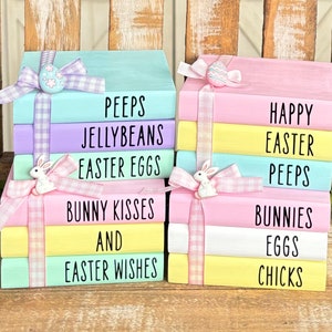 Puede incluir: Una pila de cinco libros de madera de color pastel con texto de temática de Pascua. Los libros están decorados con una cinta de cuadros blancos y rosas y una pequeña figura de conejo. El texto en los libros dice: "Peeps, Jellybeans, Easter Eggs, Bunny Kisses, And Easter Wishes" y "Happy Easter, Peeps, Bunnies, Eggs, Chicks".