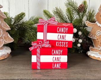 Könnte beinhalten: Ein Stapel von vier rot-weiß gestreiften Büchern mit den Worten "Candy Cane Kisses" und "Candy Cane Lane" darauf. Die Bücher sind mit einem rot-weiß gestreiften Band gebunden.