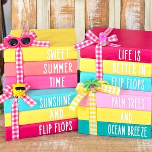 Op de afbeelding: Een stapel kleurrijke houten blokken met zomerse spreuken. De blokken zijn versierd met roze en wit gingham lint, een gele zon met een zonnebril en een roze flip-flop. De spreuken op de blokken zijn "Sweet Summer Time", "Sunshine and Flip Flops", "Life is Better in Flip Flops", "Palm Trees and Ocean Breeze".