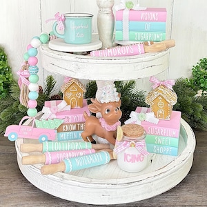 Può includere: Un vassoio a due livelli bianco decorato con una casetta di pan di zenzero, una statuetta di renna, un piccolo camion e vari mattarelli con frasi come "Sugarplums", "Lollipops" e "Nutcracker". Il vassoio presenta anche un piccolo barattolo etichettato "Icing" e una pila di libri rosa e blu con il testo "VISIONS OF SUGARPLUMS" e "SUGARPLUM SWEET SHOPPE".