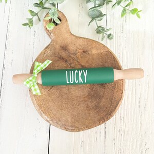 May include: Green rolling pin with a white "LUCKY" inscription on a wooden chopping board with a green and white gingham ribbon.